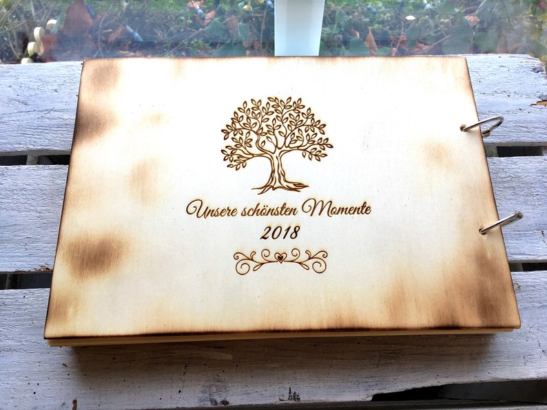 May include: A wooden photo album with a burnt wood finish. The cover features a laser-engraved design of a tree with the text "Unsere sch&ouml;nsten Momente 2018".