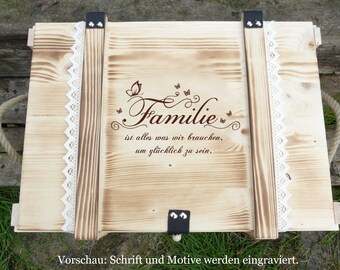 May include: A wooden box with a light brown finish, featuring a decorative inscription that reads "Familie" with a butterfly motif. The box has a white lace trim and rope handles.