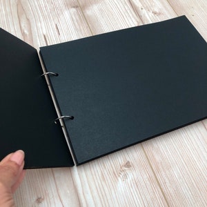 May include: A black photo album with a ring binding system. The album is open to reveal blank pages.