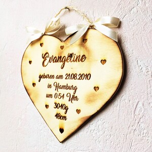 May include: A wooden heart-shaped plaque with the name "Evangeline" engraved on it. The plaque also includes the date "21.08.2010", the location "Hamburg", the time "0:54 Uhr", the weight "3040g", and the length "48cm". The plaque is decorated with small hearts and has a white ribbon attached to it.