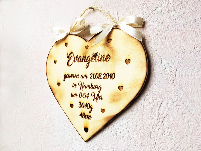 May include: A wooden heart-shaped plaque with the name "Evangeline" engraved on it. The plaque also includes the date "21.08.2010", the location "Hamburg", the time "0:54 Uhr", the weight "3040g", and the length "48cm". The plaque is decorated with small hearts and has a white ribbon attached to it.