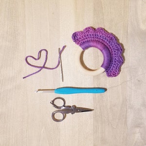 May include: A purple and white crocheted flower with a wooden ring in the center. The flower is surrounded by a crochet hook, a pair of scissors, and a length of purple yarn.