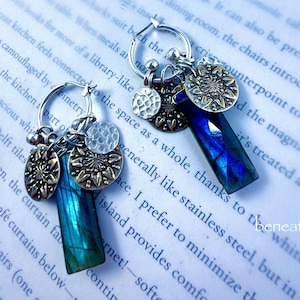 May include: A pair of hoop earrings with multiple charms. Each earring features a faceted, rectangular blue gemstone, along with silver and bronze-toned circular charms. The earrings are displayed on a background of printed text.