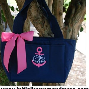 May include: A navy blue tote bag with a pink bow and an embroidered anchor with a monogram. The bag has a zippered pocket and a large pocket on the front.