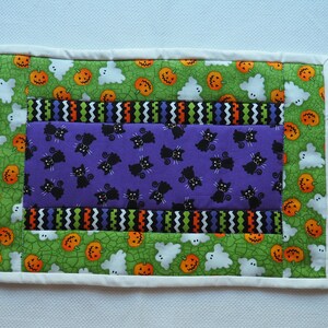 May include: A rectangular quilted fabric mat featuring a central purple panel with black cat illustrations, bordered by strips of zigzag patterns in orange, green, and black, and an outer green frame with orange pumpkins and white ghost motifs.