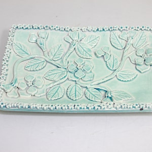 May include: A light blue ceramic dish with a raised floral design. The dish has a white border with small flower accents.