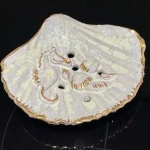 May include: A white ceramic soap dish shaped like a seashell with gold accents. The dish features two seahorse figures and three smaller seashells.