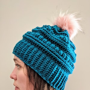 May include: A teal blue crocheted beanie with a pink faux fur pom pom on top.
