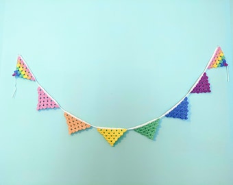 Könnte beinhalten: Eine bunte gehäkelte Girlande mit dreieckigen Flaggen in den Farben Rosa, Orange, Gelb, Grün, Blau und Lila.
