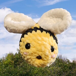 Op de afbeelding: Een geel en zwart gehaakt knuffelbeestje in de vorm van een bij met witte, pluche vleugels. De bij heeft zwarte ogen en een roze neus.