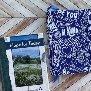 May include: A book titled "Hope for Today" next to a blue fabric book cover. The cover is decorated with white hand-drawn text and illustrations, including the words "Be Kind", "Love", and "Rainbow". A blue ribbon is attached to the cover.