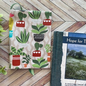 May include: A book titled "Hope for Today" by Al-Anon Family Groups, with a green cover and a photo of a field of white flowers.  The book is next to a book cover with a beige background and a pattern of potted plants in various shades of green, red, and yellow.