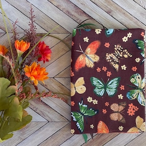 May include: A burgundy book cover featuring a colorful butterfly pattern with orange, yellow, and green butterflies. The cover also has small white and orange flower accents. A green ribbon bookmark is visible.