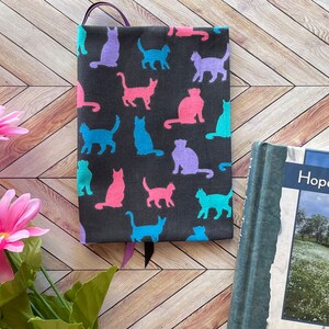 May include: A black book cover with a pattern of colorful cat silhouettes. The cover is made of fabric and has a purple ribbon bookmark. The book cover is lying on a wooden surface.