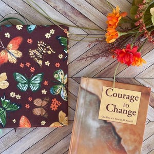 May include: A brown book cover with a butterfly pattern and a green ribbon bookmark. The book is next to a book with the words "Courage to Change" on the cover. Orange and red flowers are in the background.
