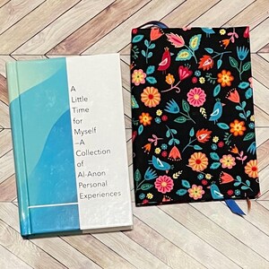 May include: Two books on a wooden surface. One book has a blue and white cover with the title "A Little Time for Myself - A Collection of Al-Anon Personal Experiences." The other book is a journal with a black cover featuring a floral and bird pattern.
