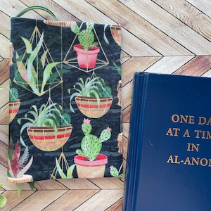 May include: A book cover with a black background and a colorful pattern of hanging plants and cacti. The book cover is made of fabric and has a loop for hanging. The book cover is next to a blue book with the title "One Day At A Time In Al-Anon" printed in gold lettering.