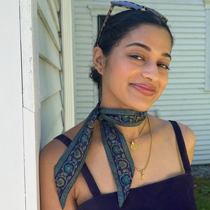 May include: A navy blue silk scarf with a floral pattern in teal, yellow, and white is tied around the neck. The scarf is paired with a gold necklace and a dark blue top. Sunglasses rest on the person's head.