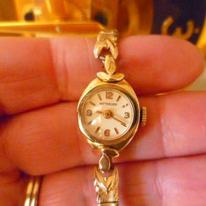 May include: A vintage gold-toned Wittnauer wristwatch. The watch has a small, oval face with a cream-colored dial and gold hands and numerals. The band is also gold-toned with a delicate, leaf-like design. The watch is held in a person's hand.