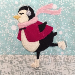 Könnte beinhalten: Ein Filz-Pinguin mit rosa Schal und Ohrenschützern beim Schlittschuhlaufen auf einem blauen und weißen Hintergrund mit Schneeflockenmuster.
