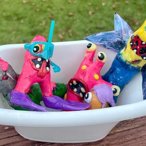 May include: A whimsical, handmade sculpture featuring colorful monster figures in a white bathtub. The monsters are various colors, including gray, pink, and yellow, with expressive eyes and mouths. The bathtub is set on a wooden surface.