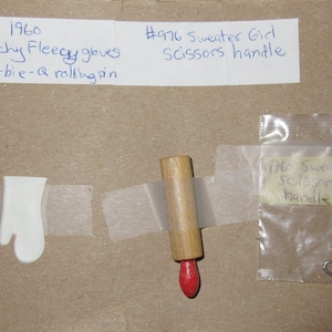 May include: An assortment of vintage Barbie doll accessories, including white gloves, a wooden rolling pin with a red tip, and a small pair of scissors. The items are labeled with handwritten notes.