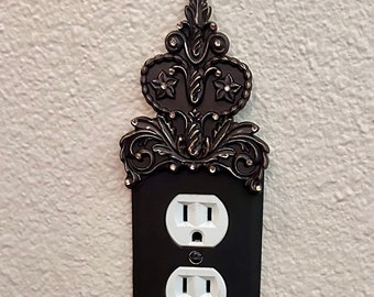 May include: A black outlet cover with a decorative ornate metal trim featuring a heart shape and rhinestones. The trim is in a dark bronze color.