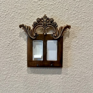 May include: A pair of light switches with a decorative, ornate, gold-colored frame. The frame has a floral design and a crown-like top.