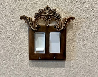 May include: A pair of light switches with a decorative, ornate, gold-colored frame. The frame has a floral design and a crown-like top.