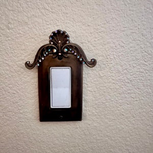 May include: A decorative light switch cover with a ornate design and rhinestones. The cover is made of metal and has a dark brown finish.