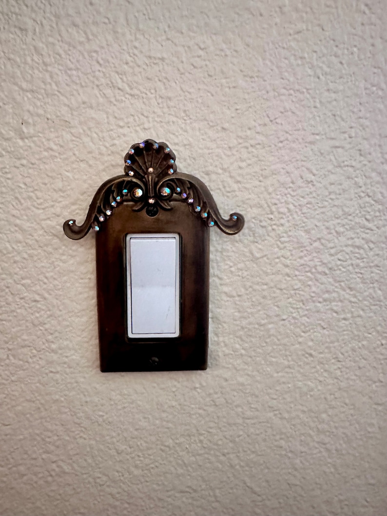 May include: A decorative light switch cover with a ornate design and rhinestones. The cover is made of metal and has a dark brown finish.