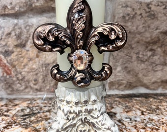 May include: A white candle holder with a decorative base and a gold fleur-de-lis design with rhinestones.