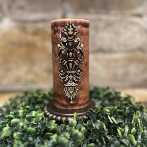 May include: A brown pillar candle with a gold decorative design. The candle is sitting on a green wreath.