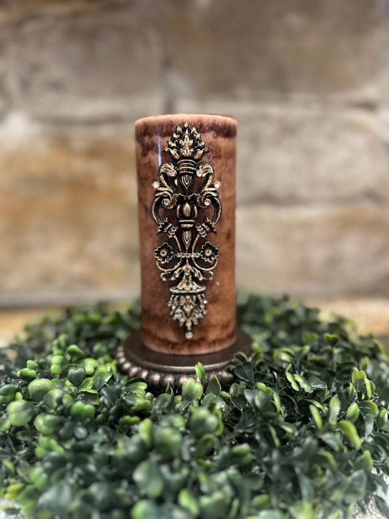 May include: A brown pillar candle with a gold decorative design. The candle is sitting on a green wreath.