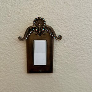May include: A decorative light switch cover with a ornate design and rhinestones. The cover is made of metal and has a light brown finish.
