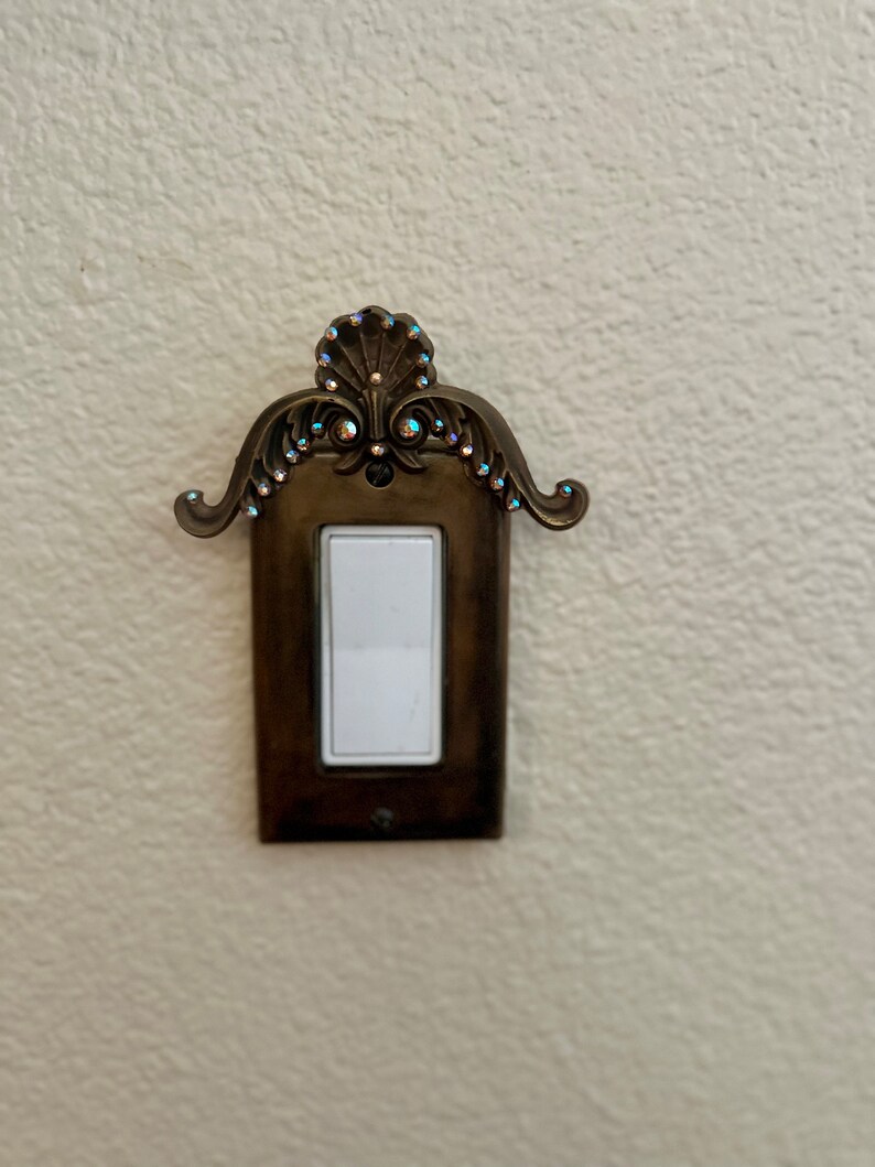 May include: A decorative light switch cover with a ornate design and rhinestones. The cover is made of metal and has a light brown finish.