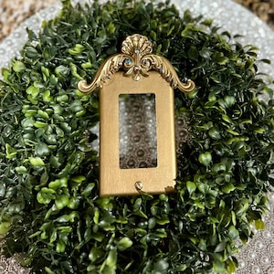 May include: Gold light switch cover with a decorative ornate frame and rhinestones. The cover is surrounded by a green wreath.