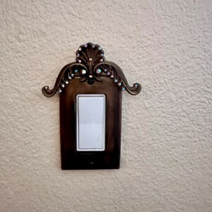 May include: A decorative light switch cover with a ornate design and rhinestones. The cover is made of metal and has a brown finish.