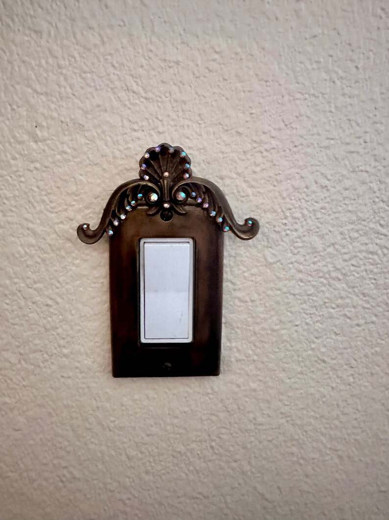 May include: A decorative light switch cover with a ornate design and rhinestones. The cover is made of metal and has a brown finish.