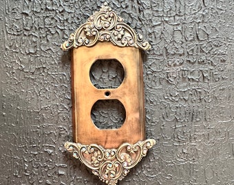 May include: A decorative outlet cover with an ornate gold frame and rhinestones. The cover is mounted on a dark textured wall.