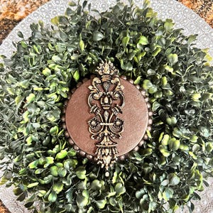 May include: A gold decorative ornament with a crown and intricate details, set against a brown background. The ornament is surrounded by a wreath of green foliage.