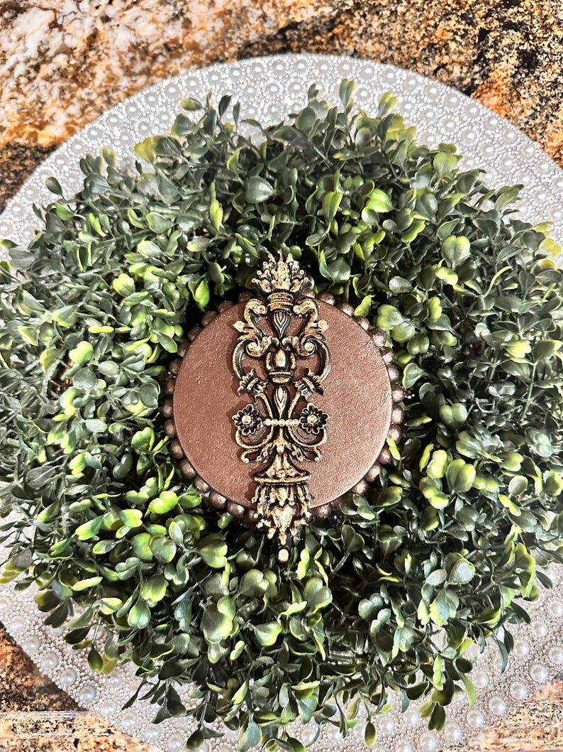 May include: A gold decorative ornament with a crown and intricate details, set against a brown background. The ornament is surrounded by a wreath of green foliage.