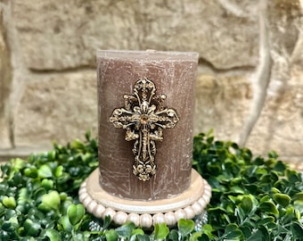 May include: A brown pillar candle with a gold cross design. The candle is sitting on a white beaded base.