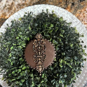 May include: A brown circular ornament with a gold-colored design featuring a stylized floral pattern. The ornament is surrounded by a wreath of green artificial foliage.