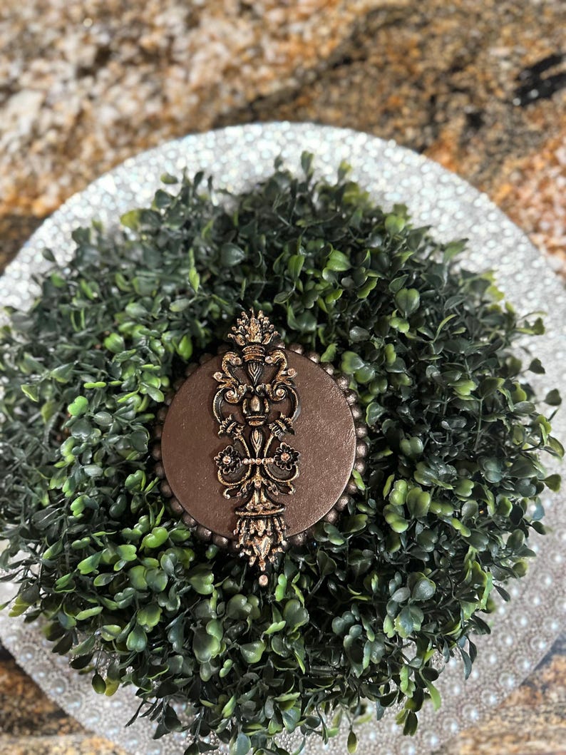 May include: A brown circular ornament with a gold-colored design featuring a stylized floral pattern. The ornament is surrounded by a wreath of green artificial foliage.