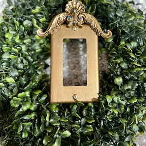 May include: A gold-colored light switch cover with a decorative, ornate design. The cover is embellished with rhinestones and has a scalloped top.