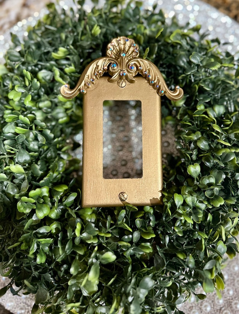 May include: A gold-colored light switch cover with a decorative, ornate design. The cover is embellished with rhinestones and has a scalloped top.