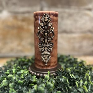May include: A brown pillar candle with a gold decorative design on the front. The candle is sitting on a green wreath.