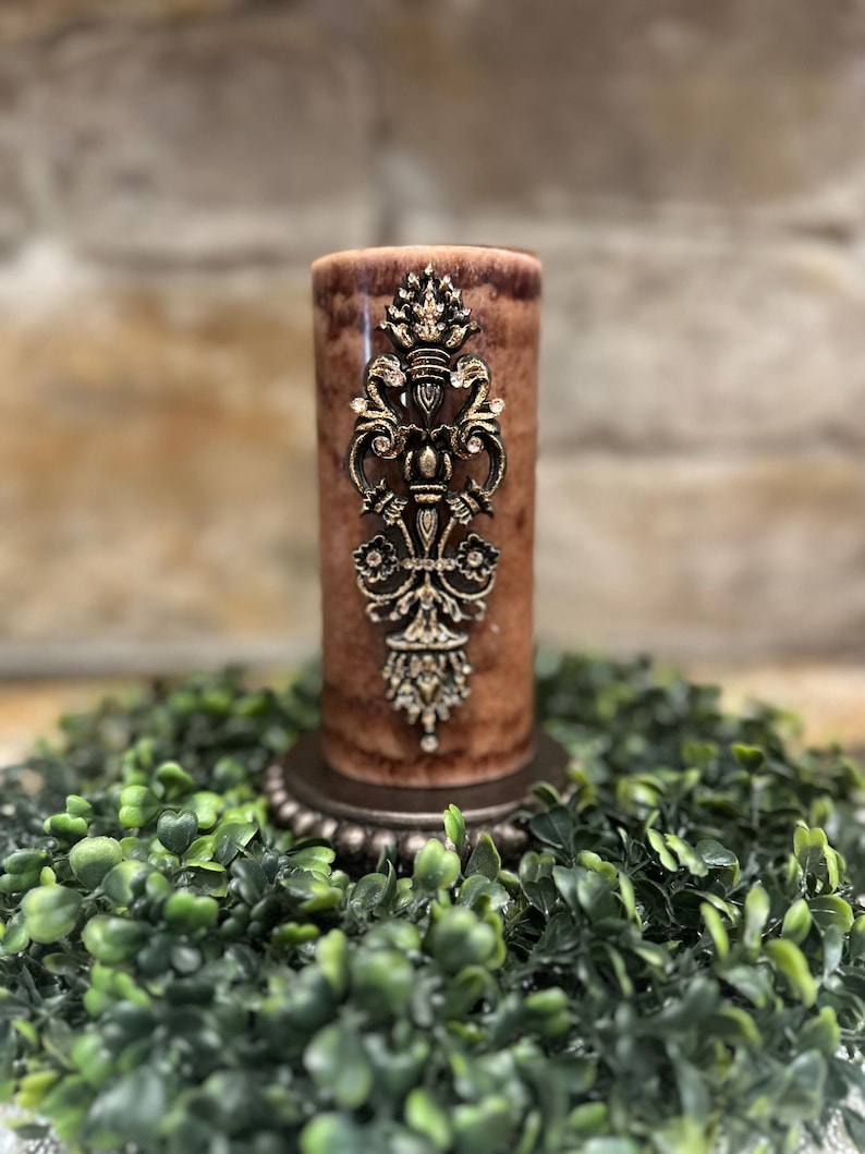 May include: A brown pillar candle with a gold decorative design on the front. The candle is sitting on a green wreath.