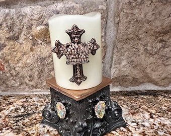 May include: A white pillar candle with a jeweled crown and cross design on a black metal stand with jeweled accents.